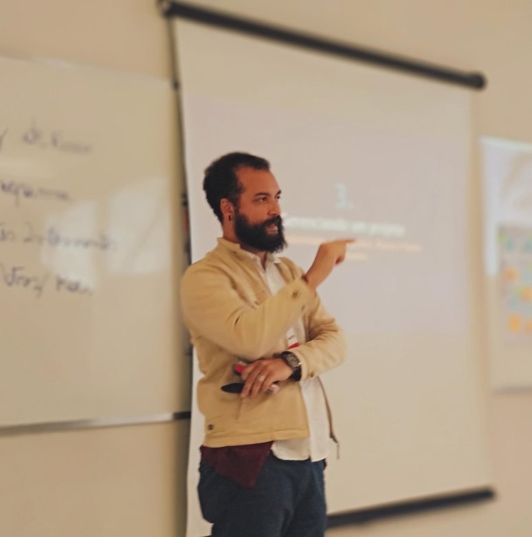 Demis em sala de aula, apontando para a projeção ao lado de um quadro branco